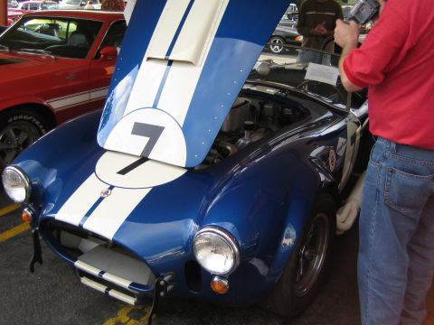 1968 Ford Mustang - A blue and white race car, possibly a Cobra, is displayed with its hood open, showing the engine. Another red car is visible in the backgro…