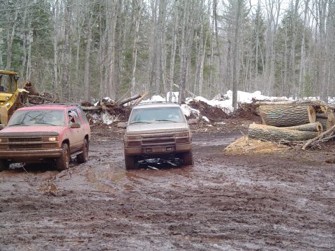 2000 Chevrolet Tahoe - Two SUVs are stuck in deep mud near cut logs and logging equipment in a wooded area.
