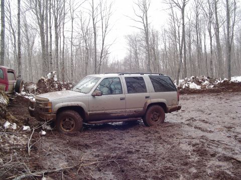 2000 Chevrolet Tahoe - This silver SUV appears to be stuck in a muddy, wooded area, with another vehicle partially visible to its left.