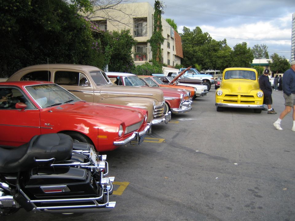 Several classic cars parked in a lot, including a red Pinto and a yellow pickup truck. A motorcycle is in the foreground