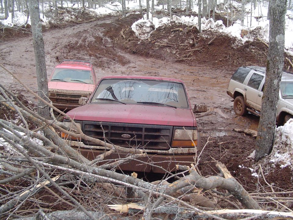 Red pickup truck stuck in mud with another vehicle behind it and a white SUV to the right