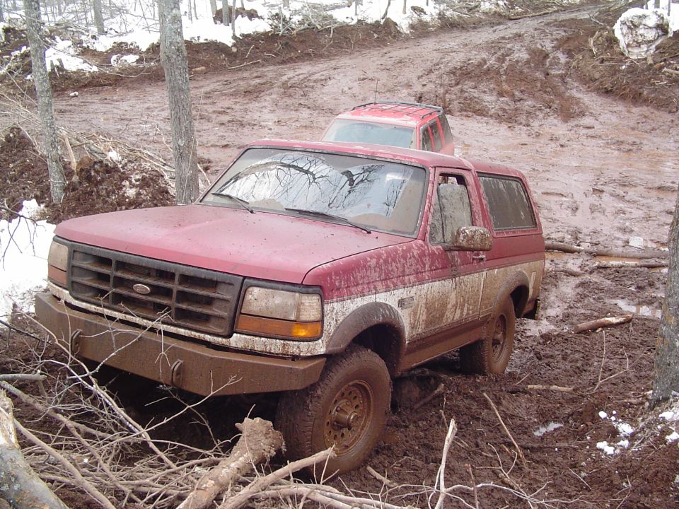 Red Ford Bronco stuck in mud with another red SUV behind it on a wooded trail
