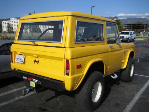 1990 Ford Bronco - Rear of a yellow early Ford Bronco showing FORD tailgate lettering, a 4x4 badge and a California license plate, parked in a lot with hills…