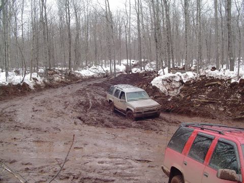 2000 Chevrolet Tahoe - Two SUVs navigate a challenging muddy trail through a forest with snow patches.