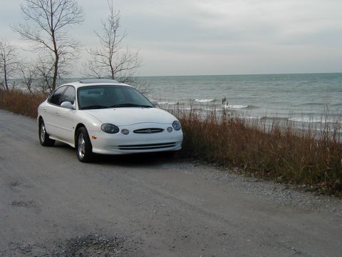 1997 Ford Taurus - A white sedan is parked on a gravel road with the ocean in the background.