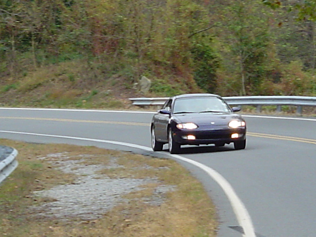 Dark blue Mazda MX-6 with headlights on rounding a curve on a rural road with guardrail behind