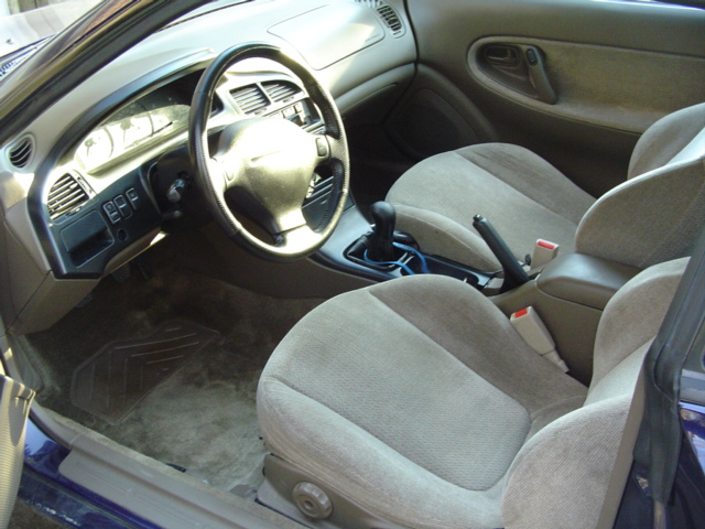 Interior of a blue coupe with tan cloth seats, manual shifter, and leather steering wheel seen through open driver door