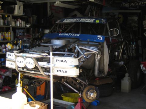 1990 Ford Bronco - number 3 off-road race truck on an exposed tube chassis with PIAA light pods and Goodyear graphics sits mid-build in a cluttered home garag…