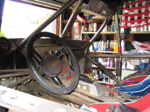 1990 Ford Bronco - Cockpit of a tube-chassis off-road race vehicle with a padded steering wheel, roll cage and gauges, backed by shelves of shop fluids.