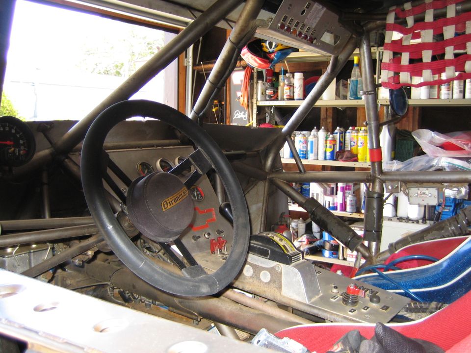 Interior of a tube-chassis race vehicle showing steering wheel, roll cage and shop shelves