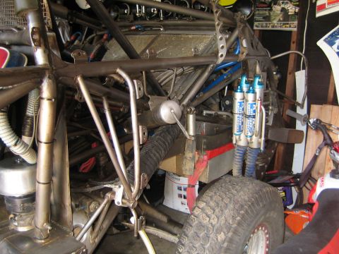 1990 Ford Bronco - bare welded tube-chassis off-road vehicle with blue bypass shocks and an exposed rear wheel sits among parts and tools in a garage.