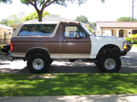 1990 Ford Bronco - Driver-side profile of a two-tone brown and white full-size Ford Bronco with a tan soft top, lifted on chrome wheels, parked under a tree.