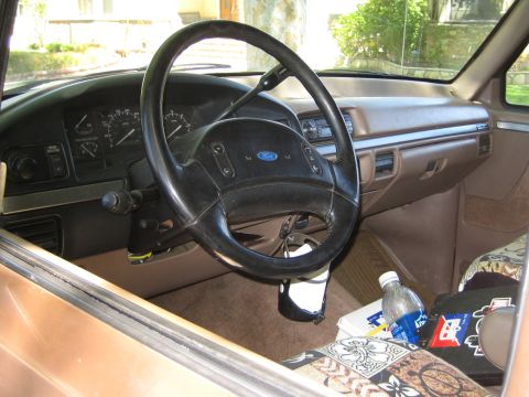 1990 Ford Bronco - Tan interior of a 1990s Ford truck or Bronco showing the steering wheel with Ford emblem, dashboard gauges and a soda bottle on the seat.