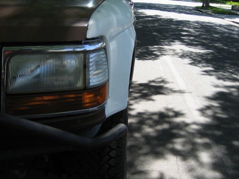 1990 Ford Bronco - Close-up of the front corner of a white full-size Ford showing a composite headlight, amber turn lens and black prerunner tube bumper on a…