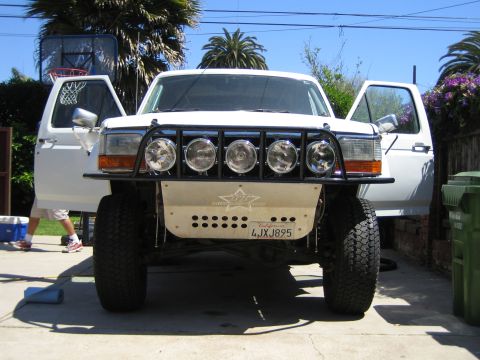 1990 Ford Bronco - Front view of a white full-size Ford Bronco with a prerunner tube bumper carrying five round driving lights, doors open in a backyard drive…