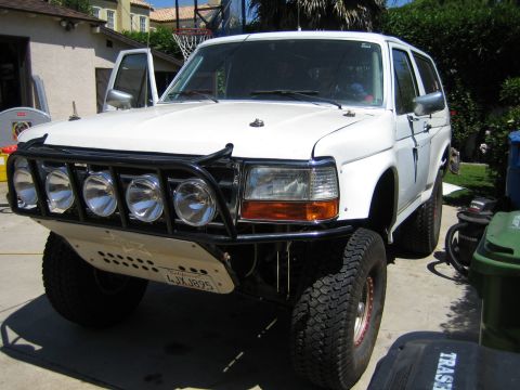 1990 Ford Bronco - white lifted full-size Ford Bronco with a prerunner tube bumper and row of driving lights sits with doors open in a residential backyard.
