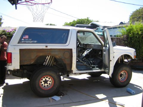 1990 Ford Bronco - Side view of a white full-size Ford Bronco mid-build, doors open showing a stripped interior and beadlock wheels, on a backyard driveway.