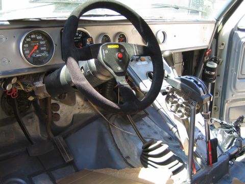 1990 Ford Bronco - Race-car cockpit featuring a Momo suede steering wheel, a large analog tachometer, pedals, and twin floor shifters.