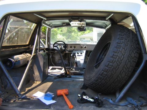 1990 Ford Bronco - Gutted vehicle interior with a roll cage, dash gauges, a spare tire standing inside, and tools on the floor.