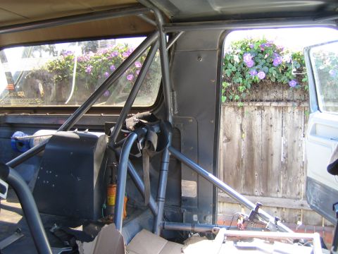 1990 Ford Bronco - Roll-cage tubing and bare interior of a vehicle, looking out a side window toward a wood fence with purple flowers.