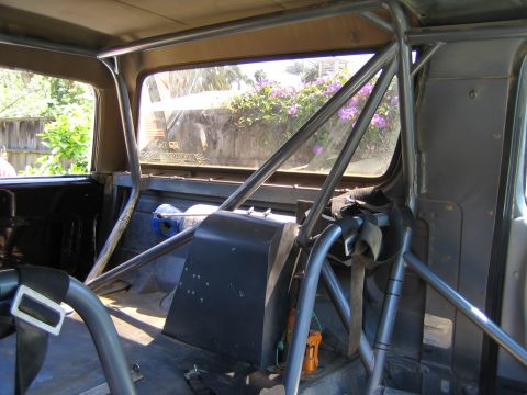 1990 Ford Bronco - Rear interior of a stripped vehicle showing roll-cage bars and a black box near the rear window with greenery outside.