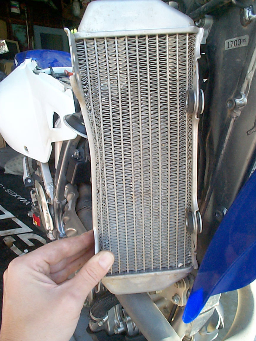 Hand holding a dirty motorcycle radiator. White plastic fairing visible to the left