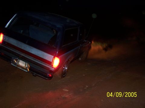 1979 Ford F-150 - A dark blue Ford Bronco is seen from the rear, kicking up dust as it drives on a dirt road at night.