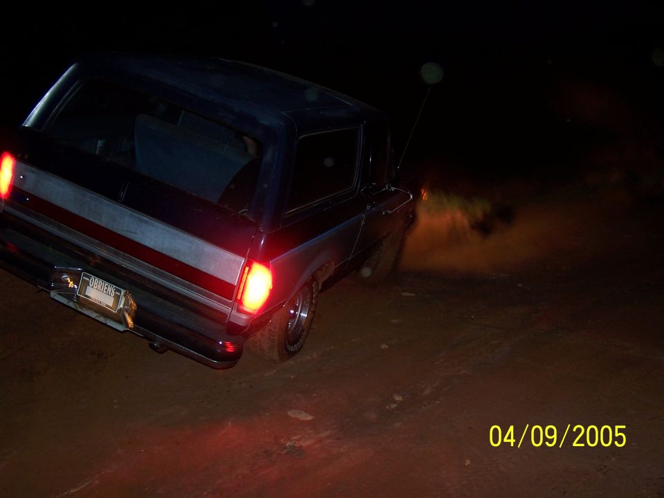 Rear view of a dark blue Ford Bronco, possibly a 1978 model, driving off-road at night. It has red taillights and a license plate that reads 'ORRIENS'