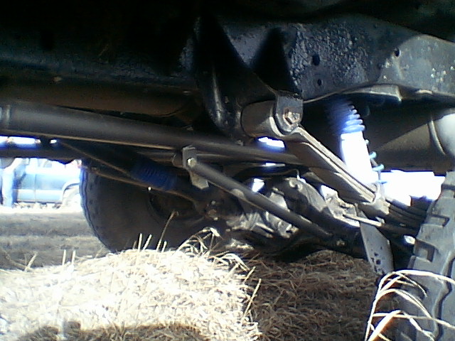 Underside of a vehicle's suspension system with blue shocks and leaf springs. Another vehicle is visible in the background