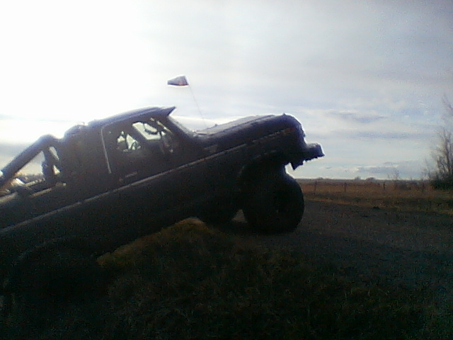 Lifted dark Ford pickup climbing a dirt embankment at dusk with whip flag, big mud tires off the ground