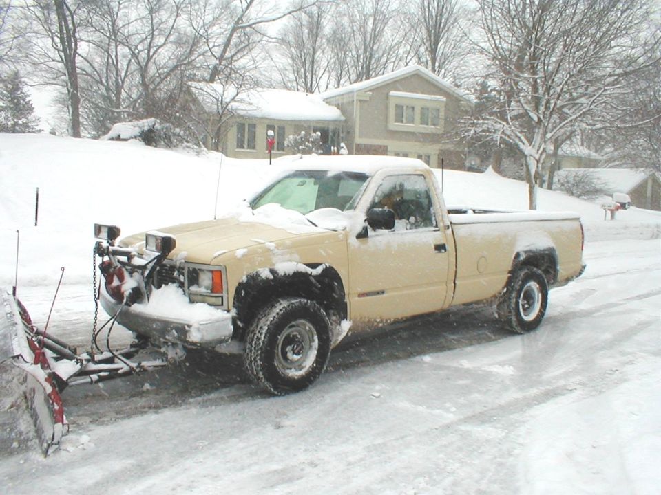 Tan single-cab GMC pickup with front snow plow on a snowy street