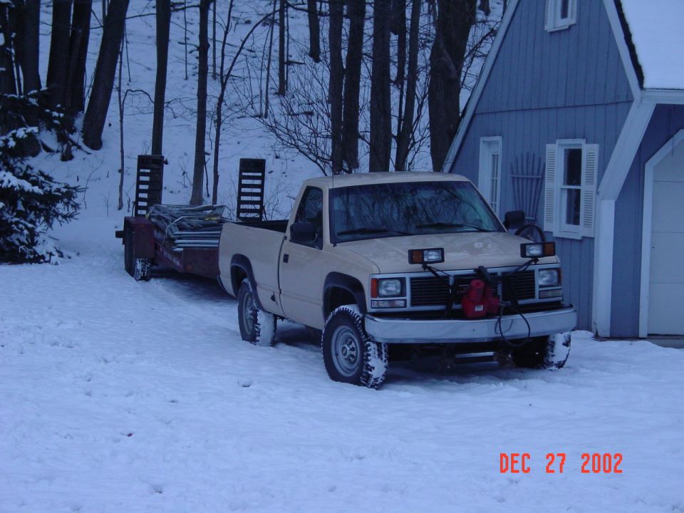 Tan GMC pickup truck with snow chains towing a flatbed trailer in snow
