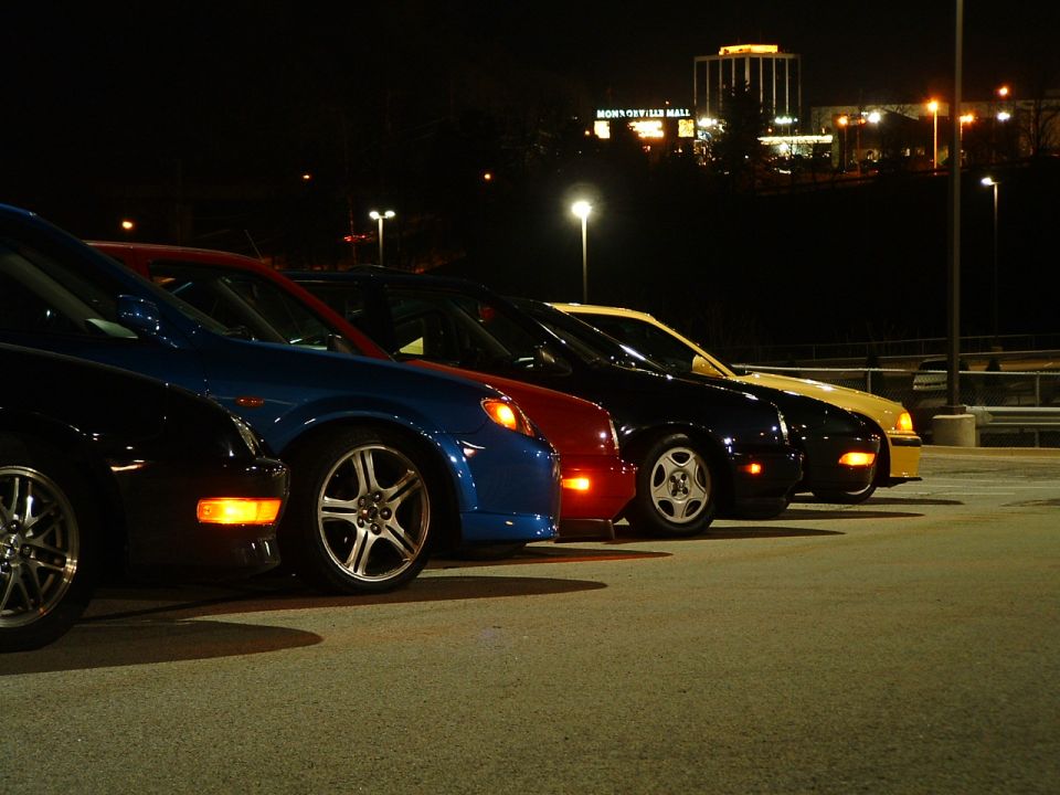Side view of multiple cars parked at night, with a mall in the background