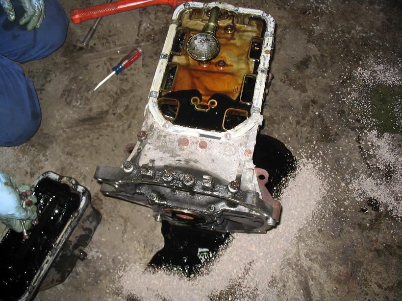 Engine oil pan with dark oil, internal components, and a screwdriver on a concrete floor