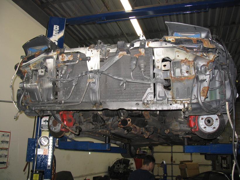 Underside of a heavily damaged vehicle's front end, suspended on a lift, showing exposed engine components, radiator, and rusted chassis