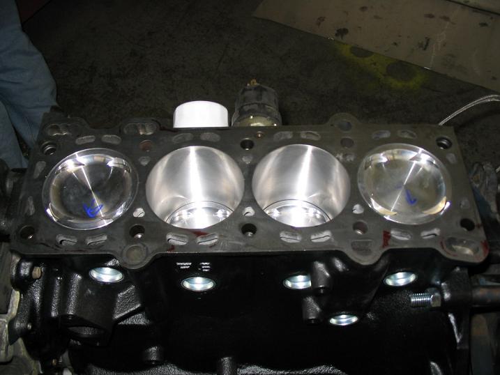 Engine block with four cylinders, two pistons visible. Gasket is on top