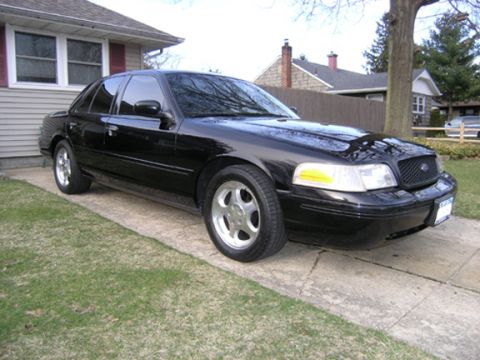 1999 Ford Crown Victoria - A black sedan is parked on a concrete driveway next to a grassy lawn.