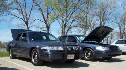 1998 Ford Crown Victoria - Two Ford Crown Victorias are parked side-by-side, one with its hood open, under a bright sky.