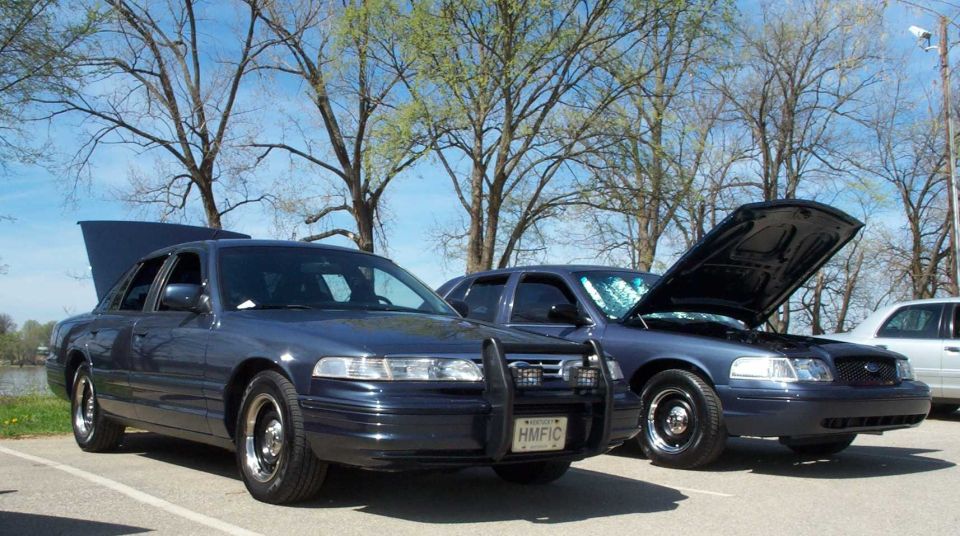 Two dark blue Ford Crown Victoria sedans at a car meet, one with trunk open and one with hood raised