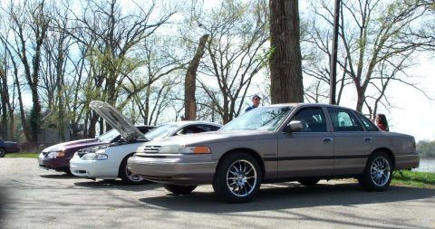 1998 Ford Crown Victoria - Several cars are parked in a line outdoors, with one car's hood open under a tree.