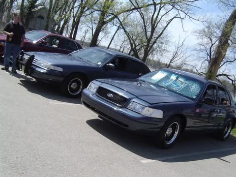 1998 Ford Crown Victoria - Two Ford Crown Victorias are parked on asphalt under trees, one with a push bar.