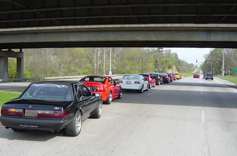 Line of Ford Mustangs parked along a roadside under a highway overpass, black Fox-body in front
