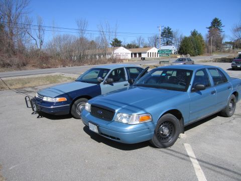 1991 Ford LTD Crown Victoria - Two blue sedans are parked in a lot on a sunny day, with one slightly in front of the other.