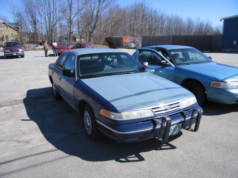 1991 Ford LTD Crown Victoria - Two police cruisers are parked in a lot on a sunny day.