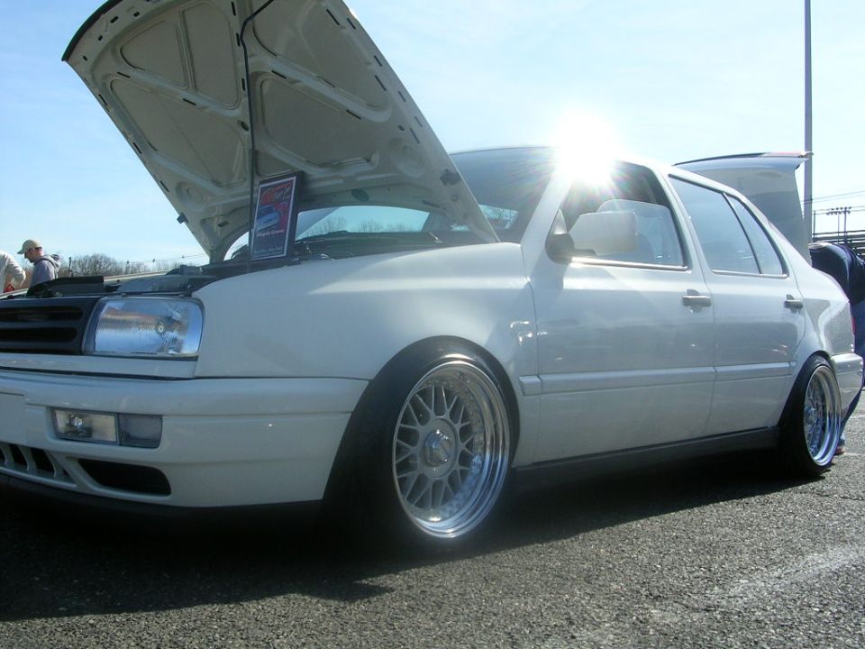 White sedan, possibly a Volkswagen Jetta, with hood open and aftermarket wheels. Sun glare is prominent