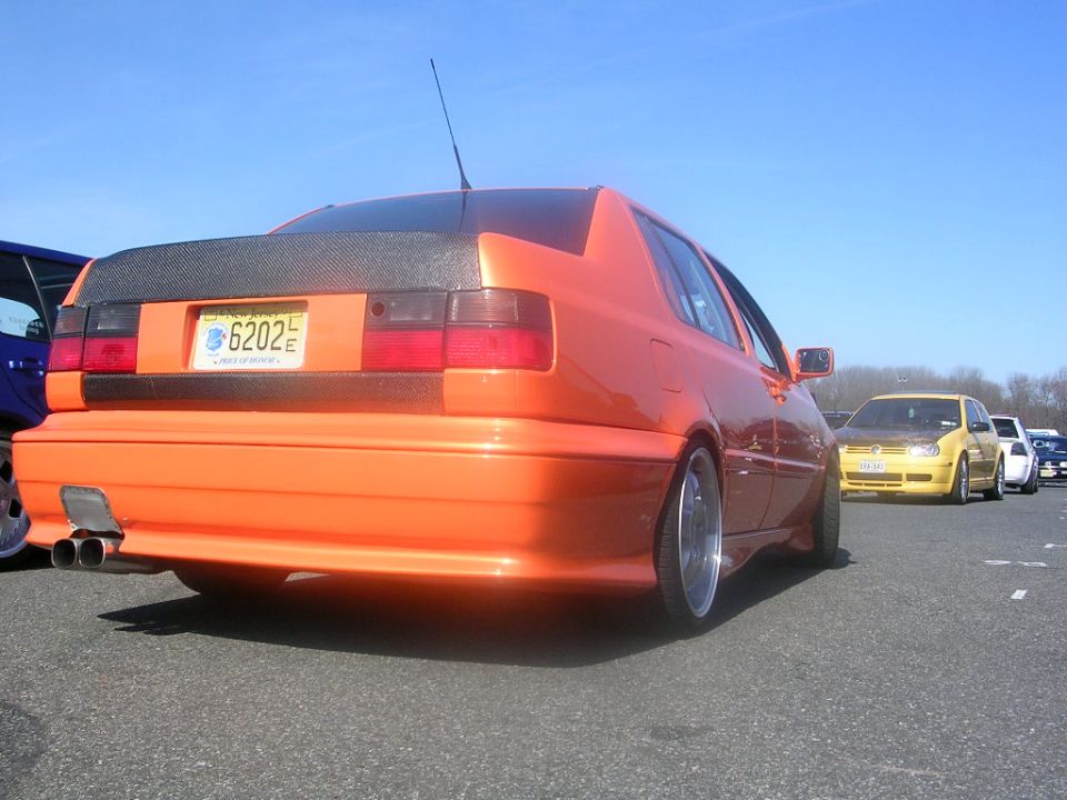 Orange Volkswagen Jetta, rear view, carbon fiber trunk lid, custom exhaust. Yellow VW in background