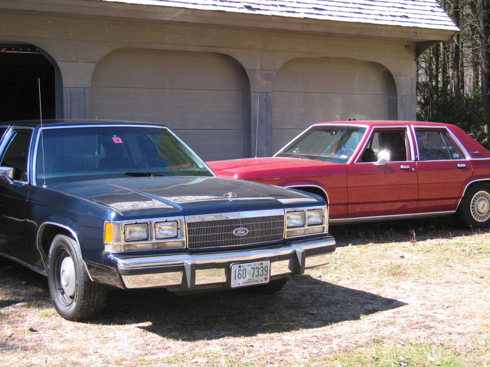Front view of a blue Ford Crown Victoria parked next to a red Ford Crown Victoria in front of a garage