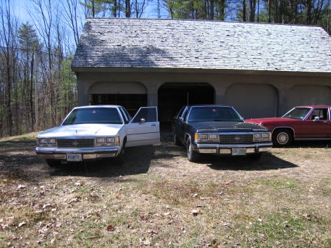 1991 Ford LTD Crown Victoria - Two Ford Crown Victorias and a red sedan are parked outside a garage on a sunny day.
