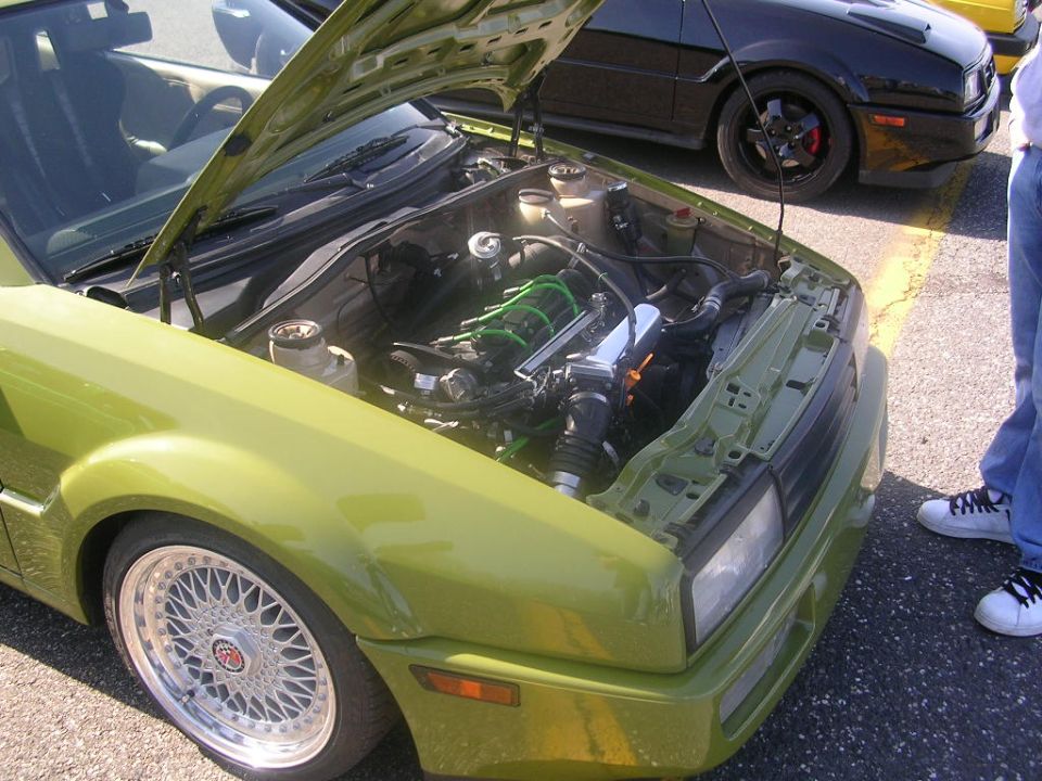 Green Volkswagen Corrado with hood open showing engine bay. Black Volkswagen in background. Person standing to the right