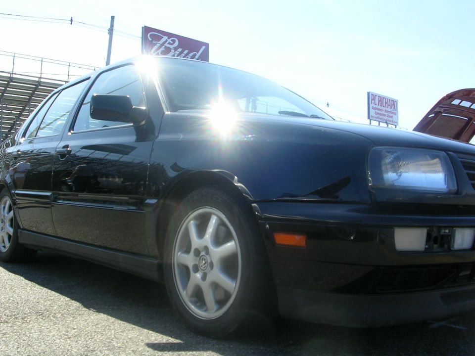 Black Volkswagen sedan, front quarter view, with a "Bud" sign and stadium seating in background